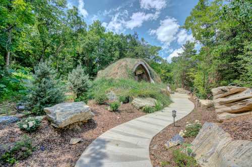 A winding path leads to a unique, grass-covered structure surrounded by trees and rocks under a blue sky.