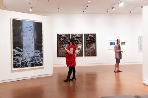 Two visitors explore an art gallery, admiring various artworks on the walls. One wears a red dress, the other a gray outfit.