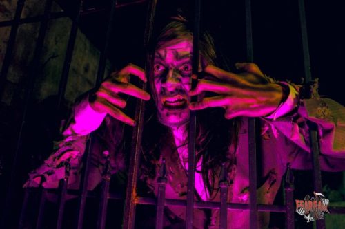 A creepy figure with long hair and tattered clothing, reaching through bars with a menacing expression in dim lighting.