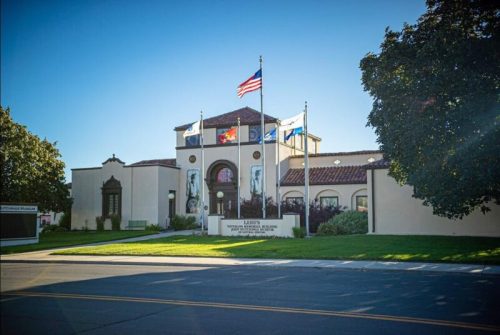 A building with a red-tiled roof, flags, and an American flag in front, set against a clear blue sky.