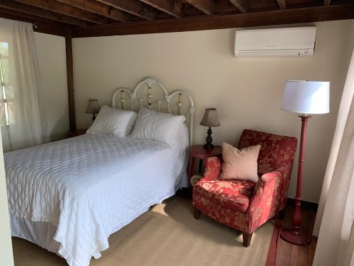 Cozy bedroom featuring a white bed, red armchair, lamps, and wooden beams in a warm, inviting atmosphere.