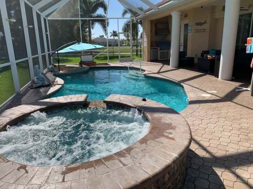 A sunny pool area featuring a blue pool, a hot tub, and a shaded lounge chair surrounded by greenery.