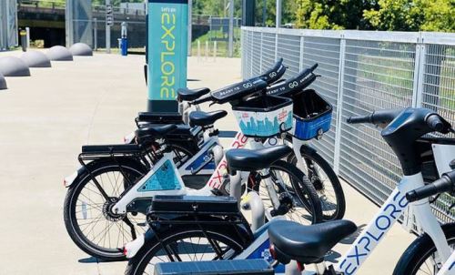 Bicycles parked at a bike-sharing station with a sign that says "Let's Explore" in a sunny outdoor setting.