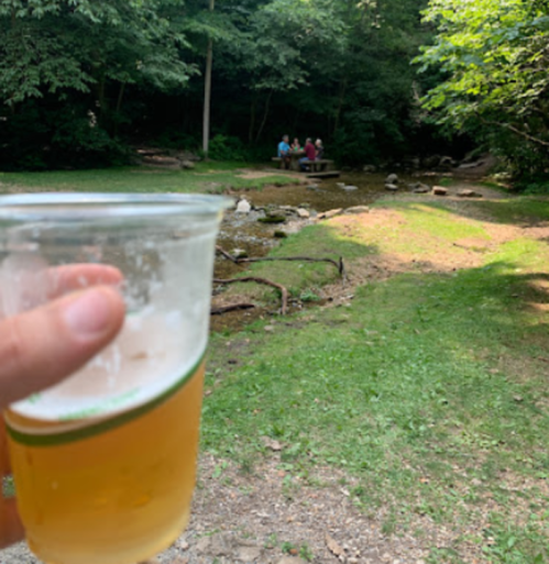 A hand holds a cup of beer in a natural setting with people sitting by a stream in the background.