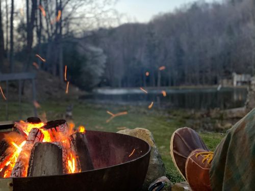 A cozy campfire with glowing embers, set by a tranquil lake surrounded by trees in the evening light.