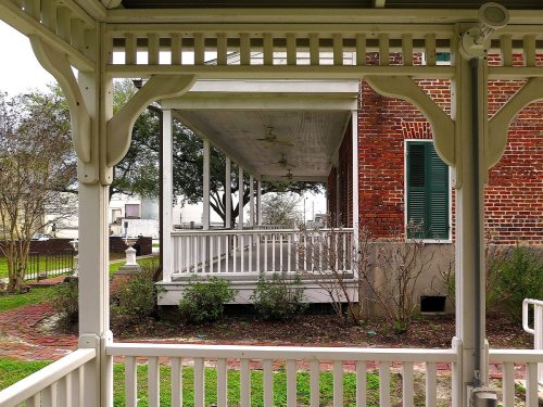 A view through a gazebo showcasing a brick building and a porch with greenery in the foreground.