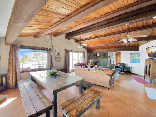 Cozy living space with wooden beams, a dining table, and a fireplace, featuring large windows and comfortable seating.