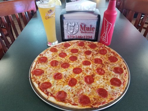 A large pepperoni pizza on a table, accompanied by a glass of beer and a red condiment bottle.