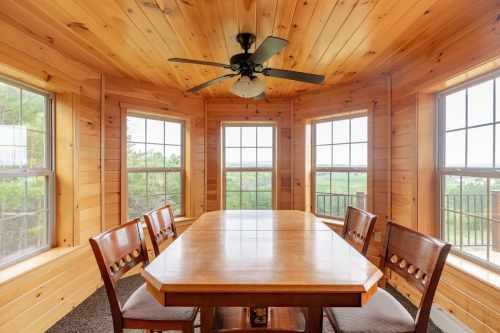 A wooden dining area with a large table and chairs, surrounded by windows offering a scenic view.