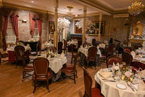 Elegant dining room with brick walls, chandeliers, and tables set for a formal meal, adorned with flowers and candles.