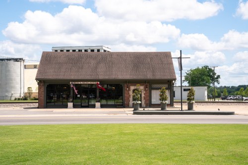 A single-story building with a sloped roof, featuring large windows and planters, set against a blue sky with clouds.