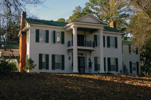 A large white colonial-style house with green shutters and a porch, surrounded by trees and fallen leaves.