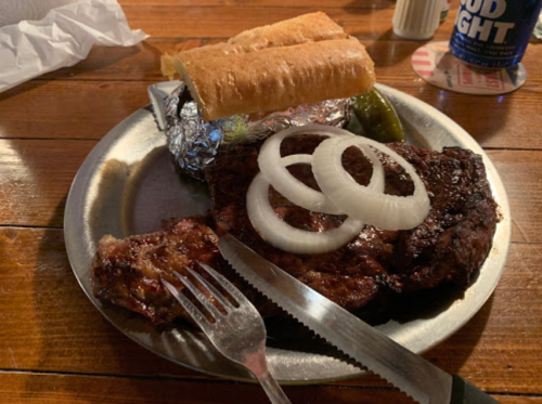 A plate with a grilled steak, sliced onions, a roll, and a foil-wrapped item, set on a wooden table.