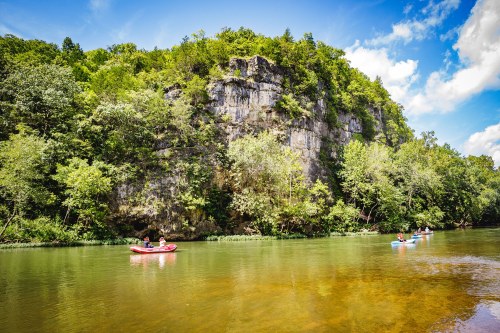 A serene river scene with kayakers paddling near a rocky cliff surrounded by lush greenery under a blue sky.