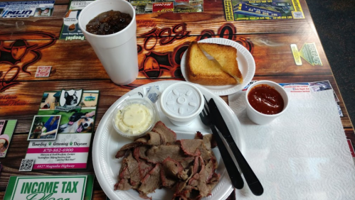 A plate of sliced meat with a side of butter, toast, and sauce, accompanied by a large drink on a colorful table.