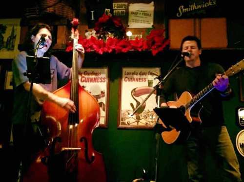 Two musicians perform on stage, one playing a double bass and the other an acoustic guitar, in a cozy pub setting.