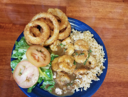 A blue plate with shrimp in sauce, rice, lettuce, tomato slices, and crispy onion rings on a wooden table.