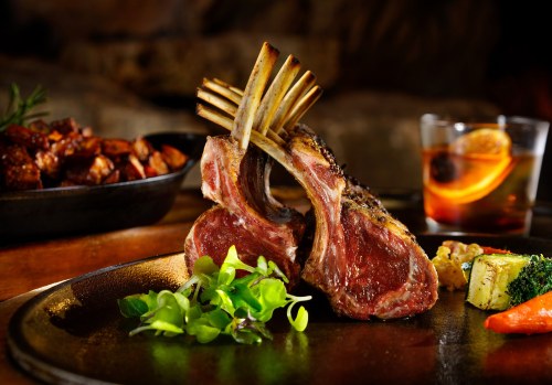 A beautifully plated rack of lamb with herbs, accompanied by roasted vegetables and a drink in the background.
