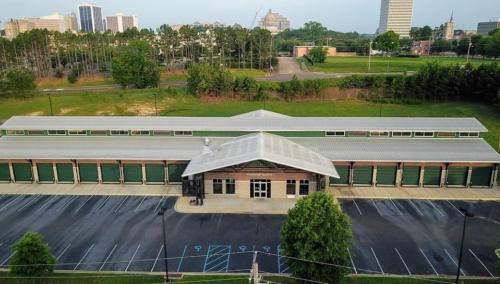 Aerial view of a storage facility with green doors, surrounded by trees and a city skyline in the background.