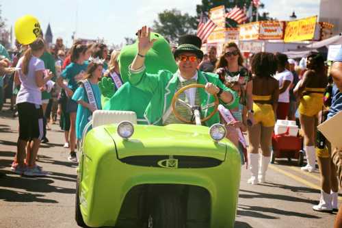 A man in a green outfit waves from a small green car, surrounded by a festive parade with colorful costumes and decorations.