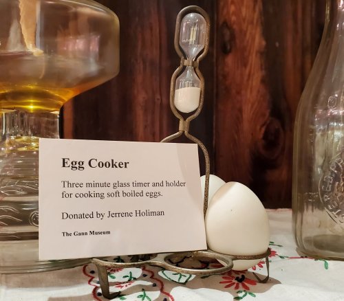 An egg cooker with a three-minute timer, displayed with a sign at the Gann Museum, alongside two eggs.