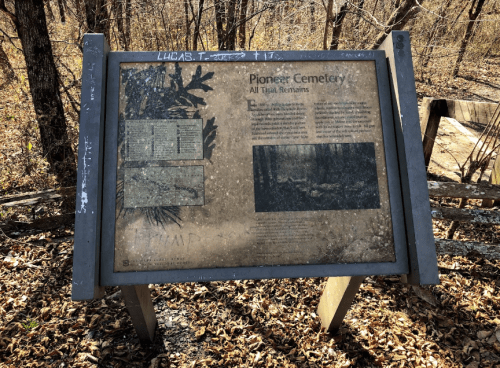 A weathered sign for Pioneer Cemetery, featuring text and images about its history, surrounded by leaf-covered ground.