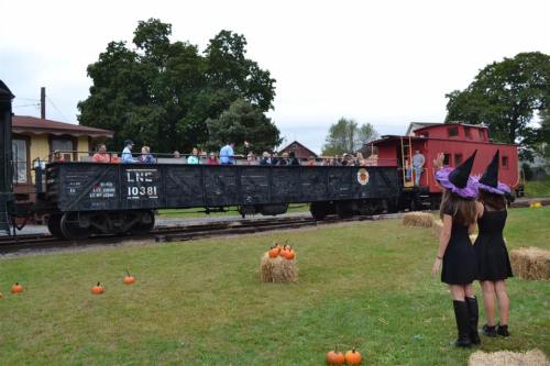 A train car filled with people at a fall festival, with pumpkins and hay bales in the foreground. Two girls in witch costumes watch.