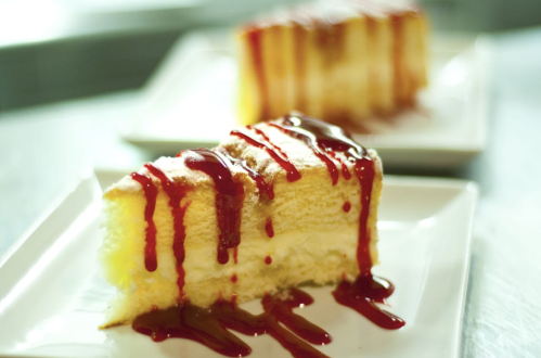 A slice of cake drizzled with red sauce, served on a white plate, with another slice in the background.