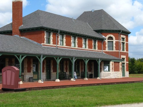Historic brick train station with a gray roof, green accents, and a spacious platform surrounded by grass.