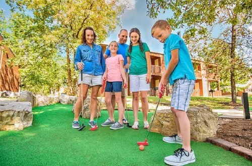 A family enjoys mini-golf outdoors, with a boy preparing to putt while others watch and smile.