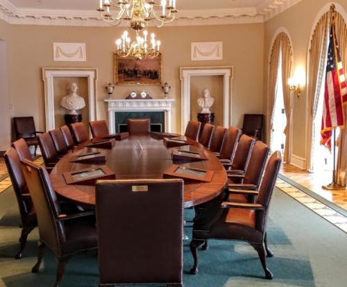A large, formal conference room with a round table, leather chairs, and American flags, featuring classical decor.