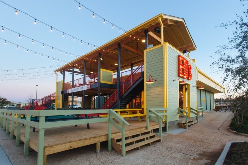 A colorful two-story building with outdoor seating and string lights, set against a clear sky.