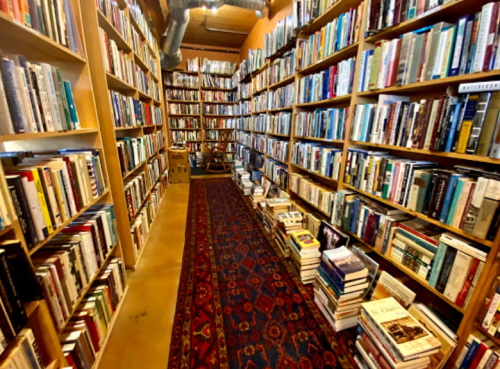 A cozy bookstore aisle lined with tall shelves filled with books, featuring a patterned rug and stacks of books on the floor.