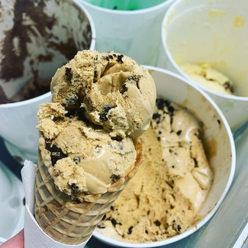 A close-up of a cone with a scoop of coffee-flavored ice cream, with empty ice cream containers in the background.
