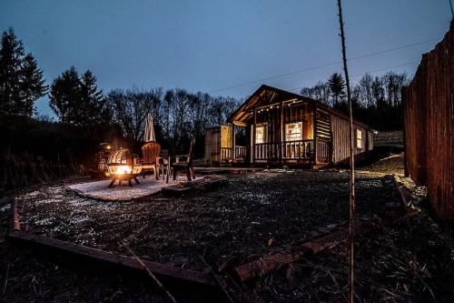 A cozy cabin at dusk, with a fire pit and chairs in front, surrounded by trees and a peaceful outdoor setting.
