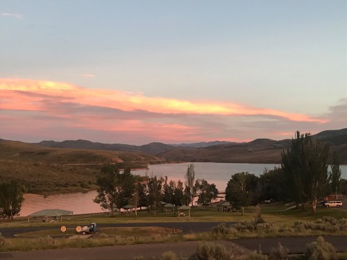 A serene landscape at sunset, featuring a lake, rolling hills, and trees under a colorful sky.