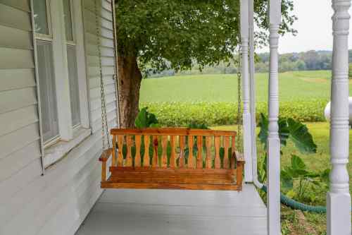 A wooden swing on a porch, overlooking a green field and trees in the background.