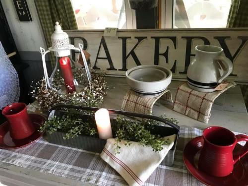 A cozy bakery display with red mugs, a candle, greenery, and rustic pottery on a table with a sign in the background.