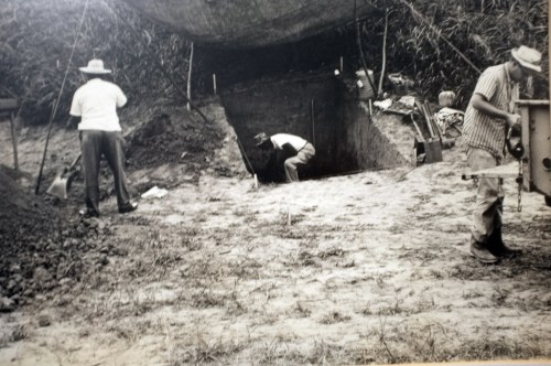 Three men working at a dig site, with one man entering a tunnel and two others digging nearby under a canopy.