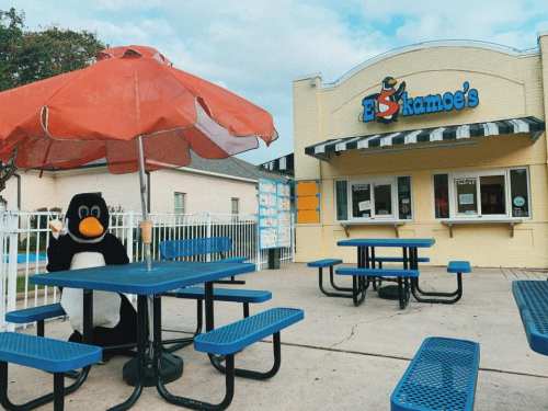 A penguin mascot waves from under a red umbrella at a colorful outdoor eatery with blue picnic tables.