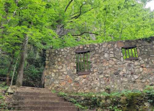 Stone wall with barred windows surrounded by lush green trees and a stone staircase leading up.