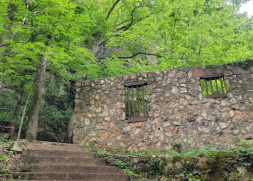 Stone wall with barred windows surrounded by lush green trees and a stone staircase leading up.
