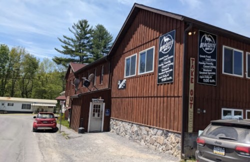 A rustic wooden building with signs for a restaurant, surrounded by trees and parked cars on a sunny day.