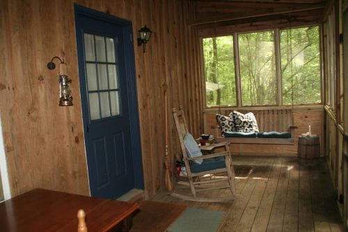 A cozy wooden porch with a blue door, rocking chair, and a cushioned bench, surrounded by greenery.