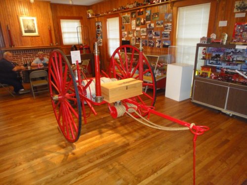 A vintage red fire cart displayed in a wooden room with photos and memorabilia in the background.