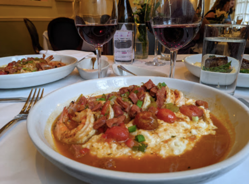 A bowl of shrimp and grits topped with tomatoes and green onions, with glasses of red wine in the background.