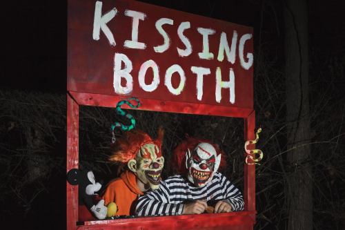 Two clowns with eerie makeup sit in a red "Kissing Booth" at night, surrounded by dark trees and playful decorations.