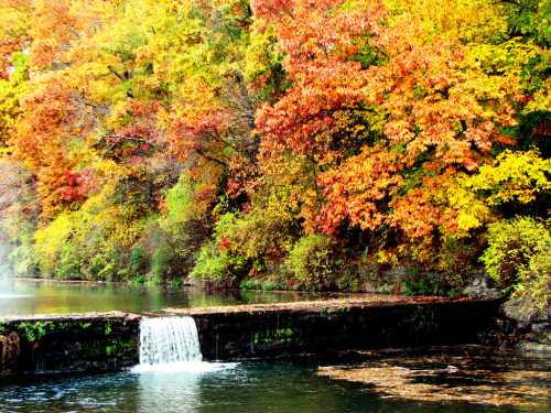 A serene waterfall flows into a calm river, surrounded by vibrant autumn foliage in shades of red, orange, and yellow.