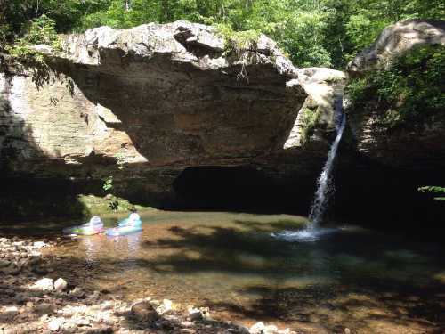 A serene natural scene featuring a small waterfall cascading into a clear pool, with two people floating on inflatable rafts.