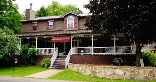 A large brown house with a porch, red shutters, and a stone pathway, surrounded by greenery.